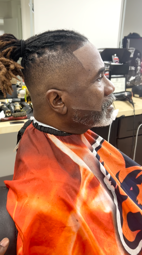 A high fade with a sharp line-up cut around locs tied in a top knot, with the beard shaped to match