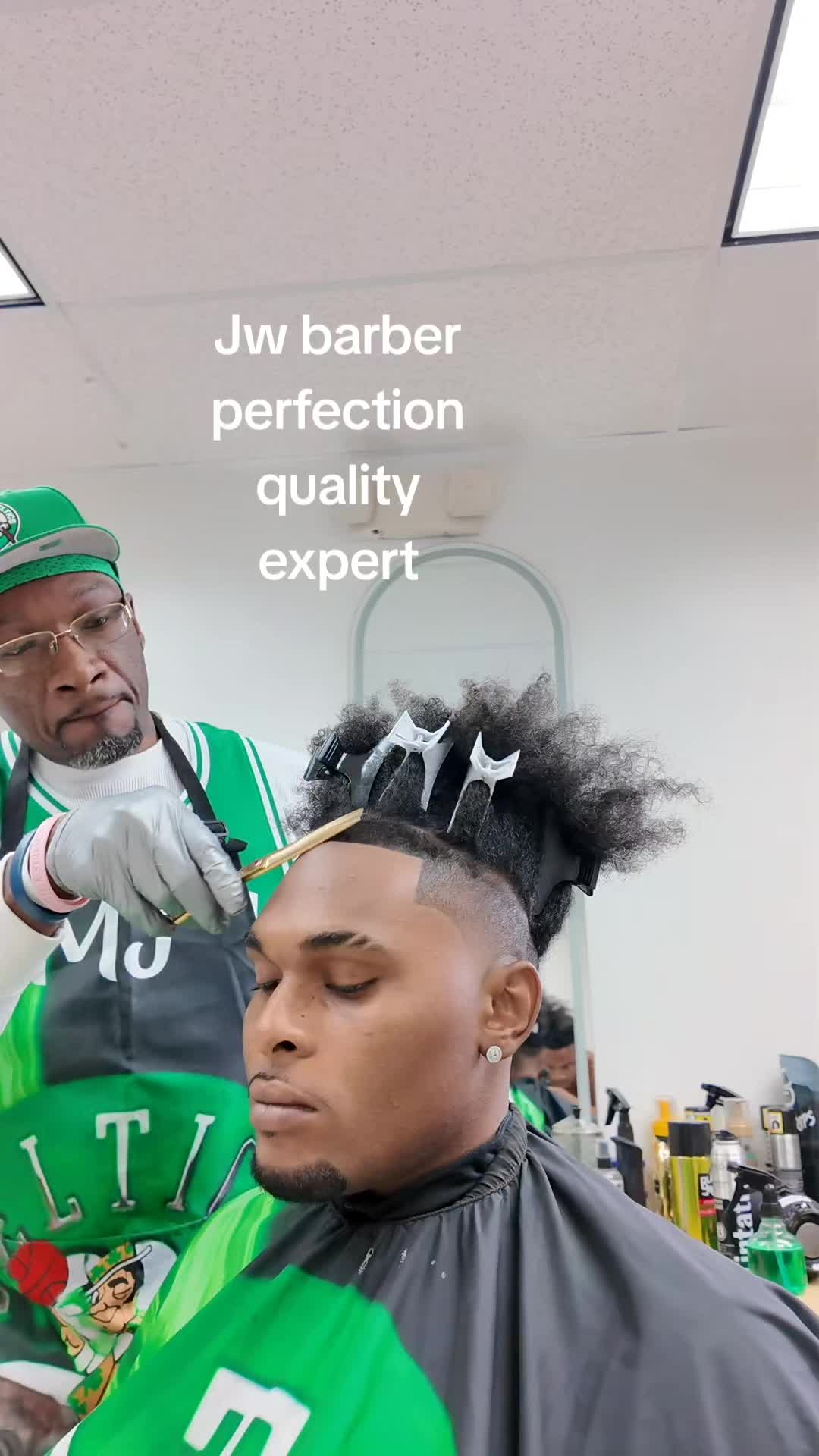 A barber in a green cap and apron shaping a client's hairline with a straight razor in the Jalia Walda barber chair